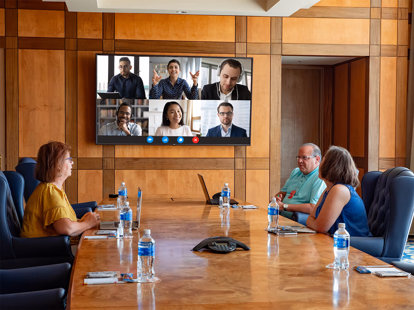 A group of people are gathered around a conference table, with a large video screen displaying several individuals in a virtual meeting at Moody Gardens Hotel's Executive Board Room in Galveston, Texas
