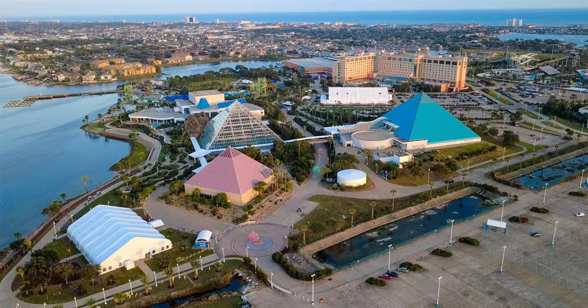 Aerial view of Moody Gardens in Galveston, Texas, showing multiple pyramid-shaped buildings, surrounding greenery, parking lots, and nearby bodies of water with the cityscape and ocean in the background.