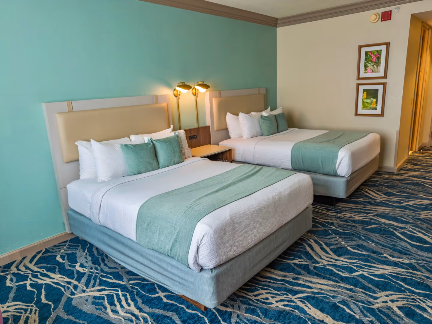 Hotel room with two queen beds dressed in white and teal linens, a shared nightstand, teal accent wall, and patterned blue carpet.