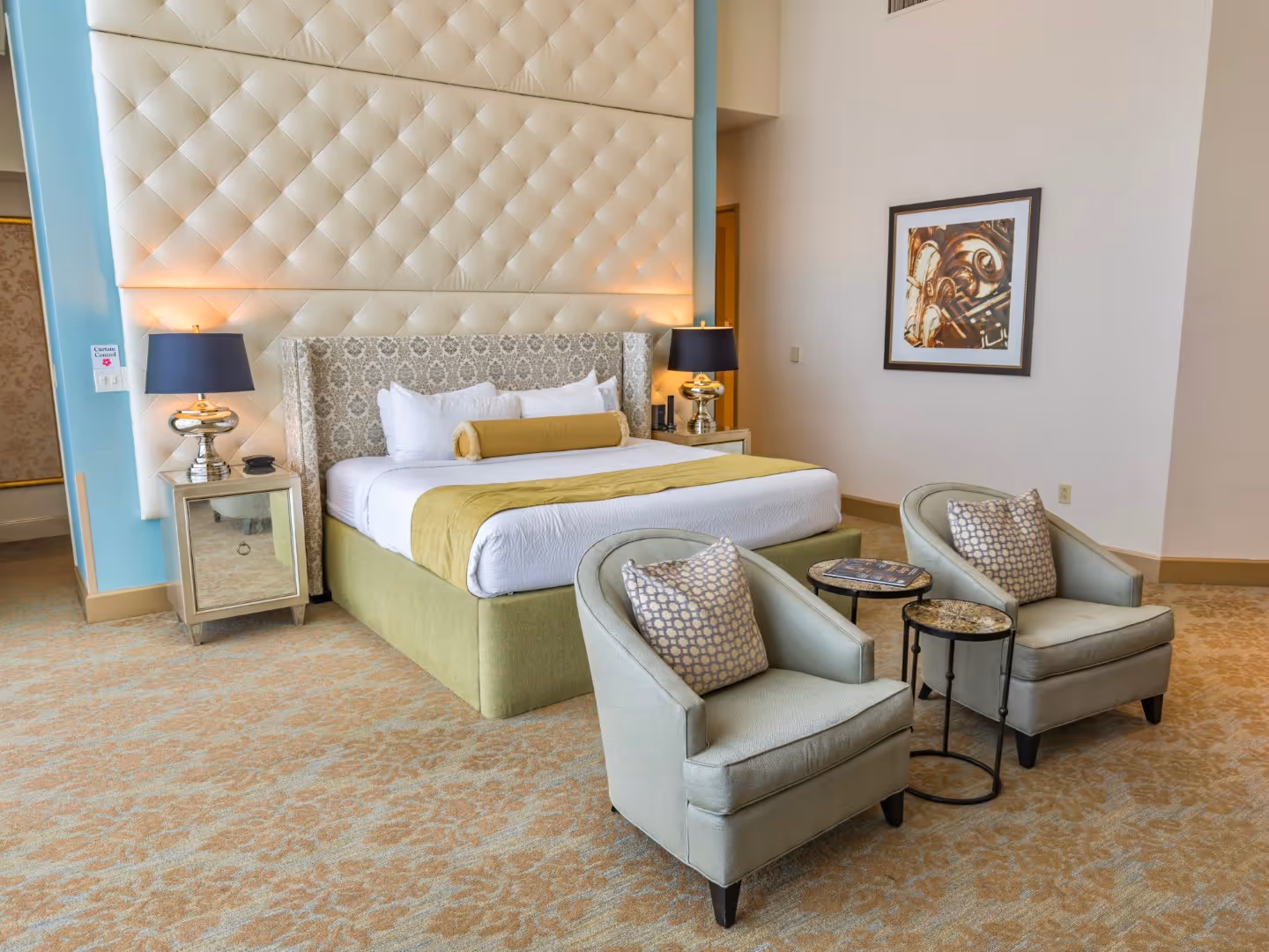 Luxury hotel room with a large bed featuring green and white bedding, two armchairs with patterned pillows, and two side tables with lamps.