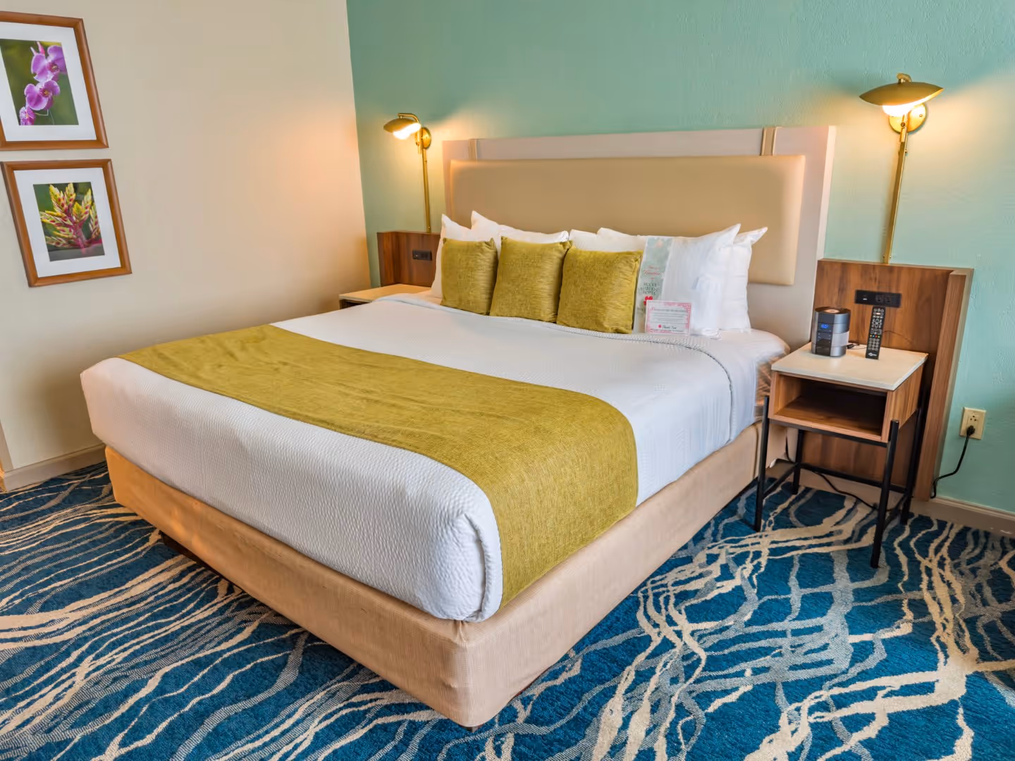 King-size bed with white bedding, olive green runner and pillows, two framed floral pictures on wall, and bedside tables with lamps and remote control.