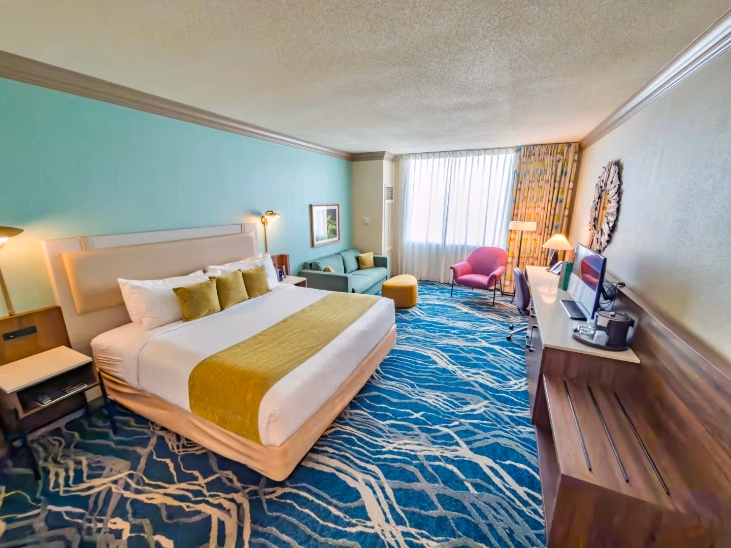 Hotel room with a king-size bed, light green accent wall, blue patterned carpet, turquoise sofa, purple armchair, and a desk with a TV and lamp.