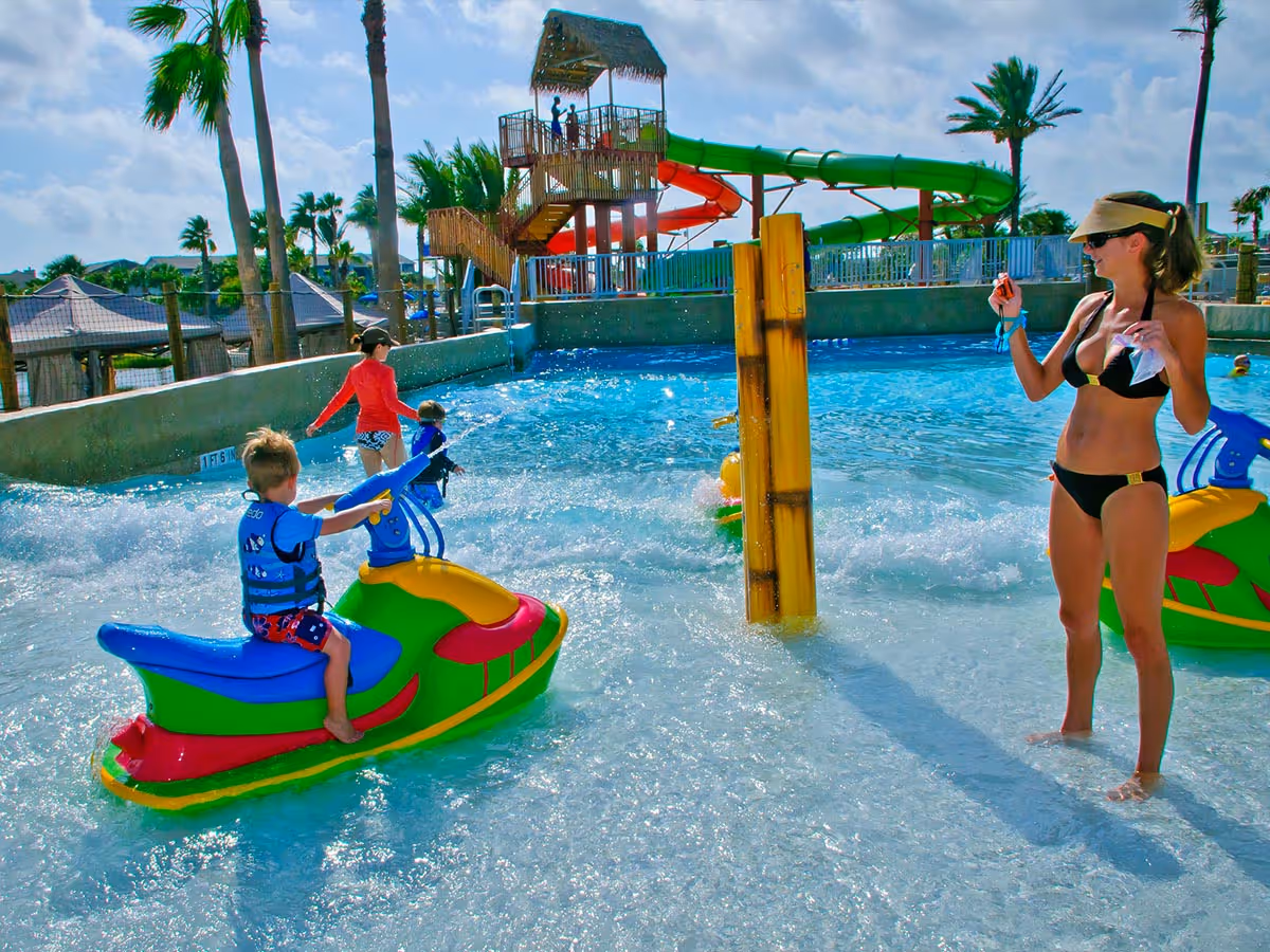 Children playing on colorful water bikes in a shallow wave pool with a woman standing nearby, a water slide and palm trees in the background.