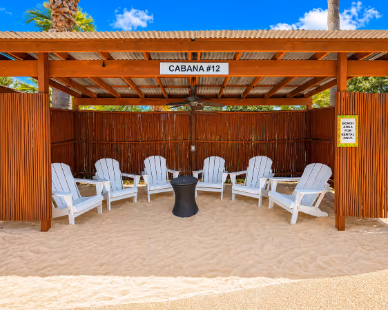 Outdoor cabana labeled 'CABANA #12' with six white Adirondack chairs arranged around a small black table on sand.