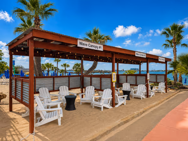 Three wave canopy huts with white Adirondack chairs on sandy ground under a blue sky with palm trees nearby.