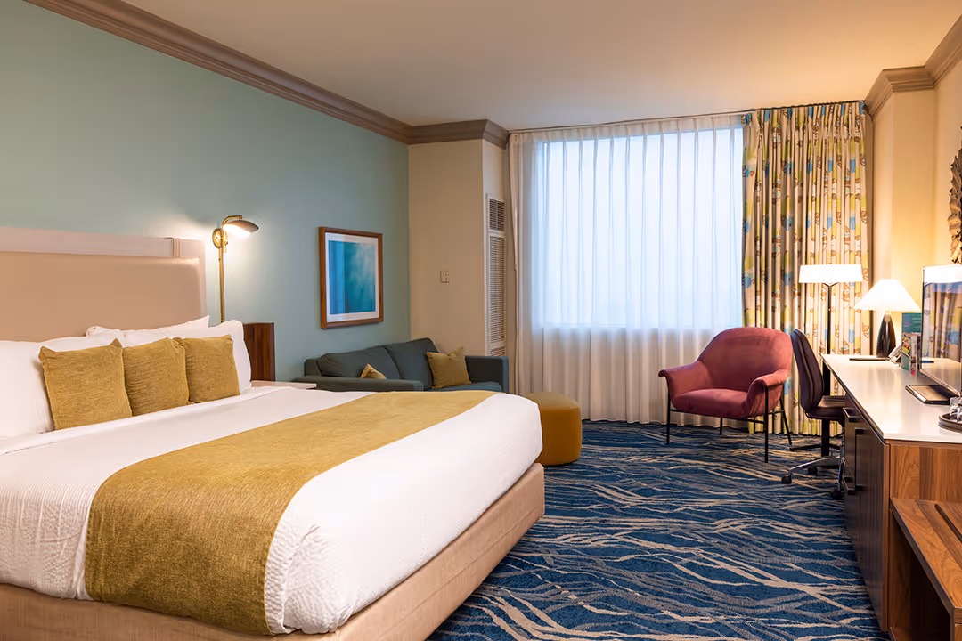 Hotel room with seating area including blue and beige armchairs, a blue sofa, a desk with a chair, and glass doors leading to a balcony with outdoor chairs.