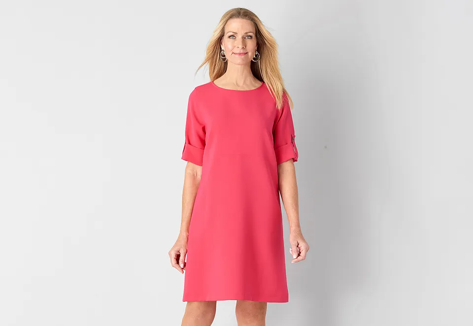 Woman with long blonde hair wearing a coral pink knee-length dress with short sleeves and hoop earrings, standing against a light gray background.