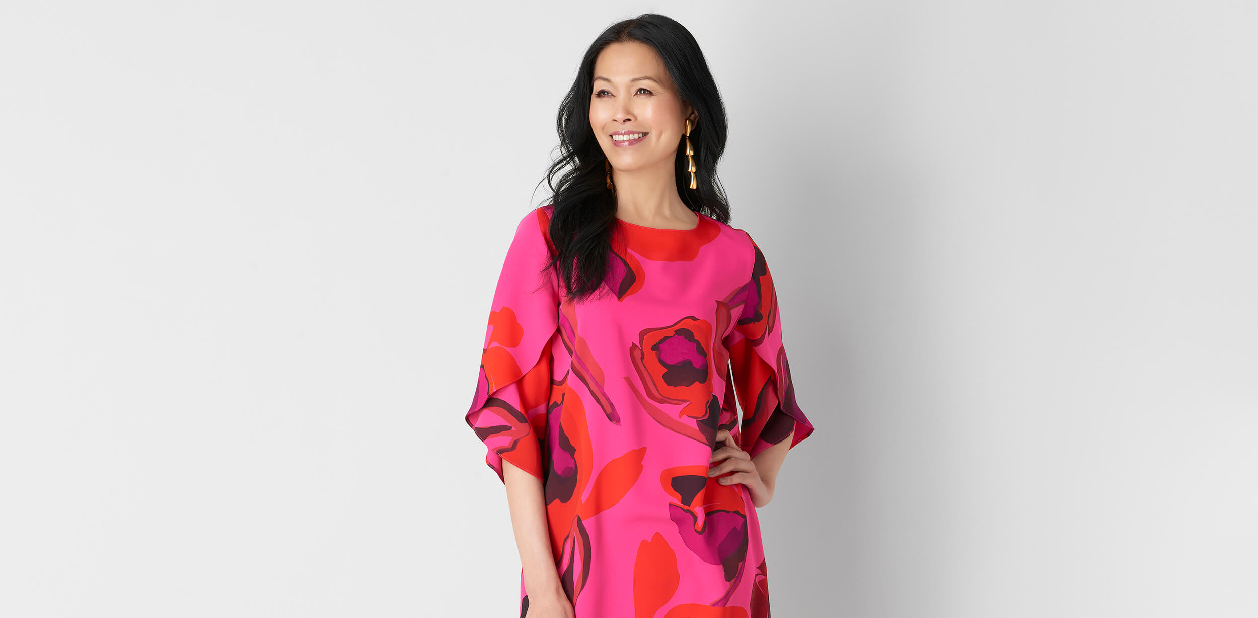 Smiling woman with wavy brown hair wearing a black and white floral print dress with three-quarter sleeves against a light gray background.