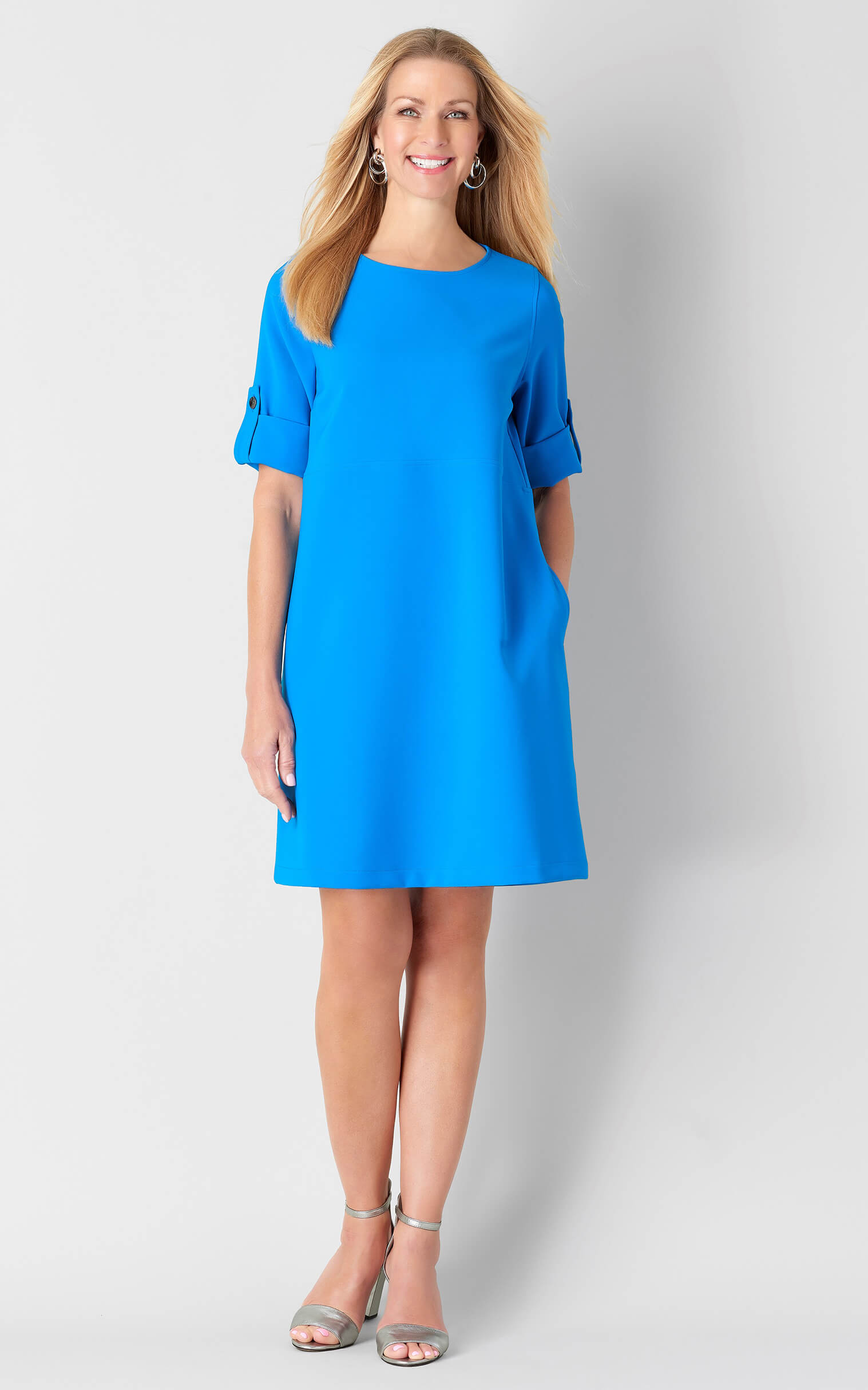 Smiling woman with long blonde hair wearing a bright blue knee-length dress and silver ankle-strap heels against a white background.