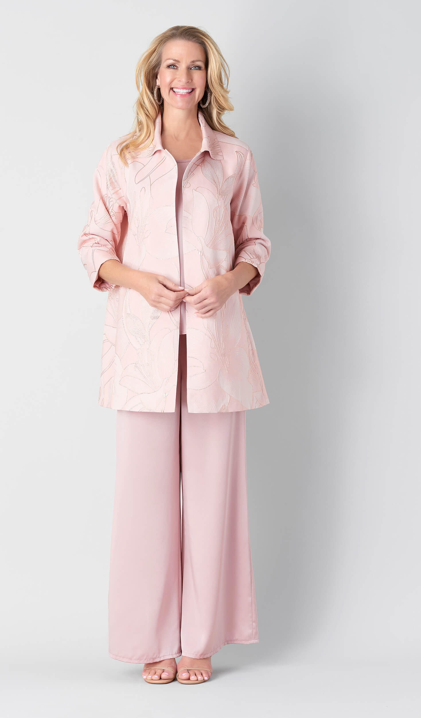 Smiling woman wearing a light pink floral-patterned jacket with wide-leg pink pants and open-toe sandals against a plain background.