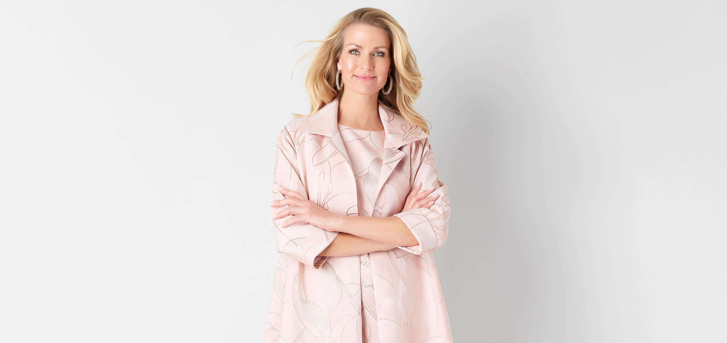 Smiling woman with blonde hair wearing a light pink floral patterned jacket and dress, standing with arms crossed against a white background.