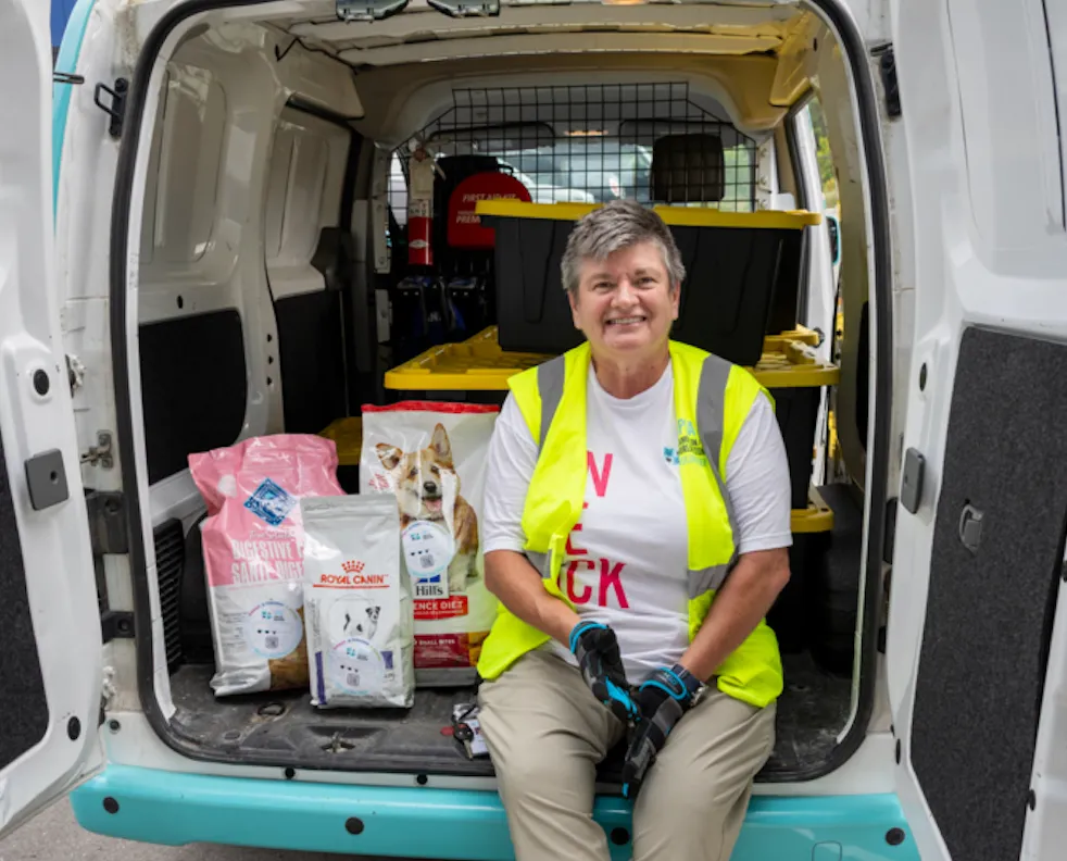 Smiling person in a yellow safety vest sitting in the back of a delivery van with bags of dog food and storage bins.
