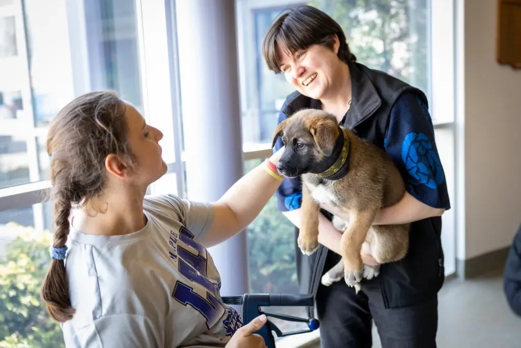 A smiling young woman holding a puppy interacts with another woman in a wheelchair near a window.