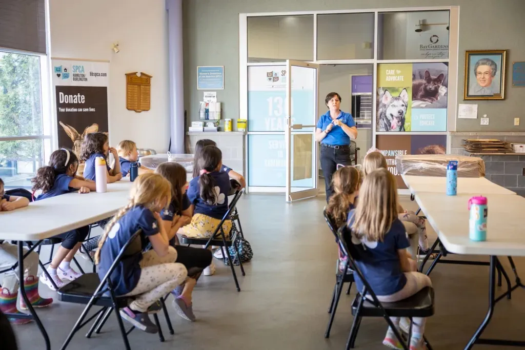 Group of children seated at tables attentively listening to a woman speaking in a classroom or community center.