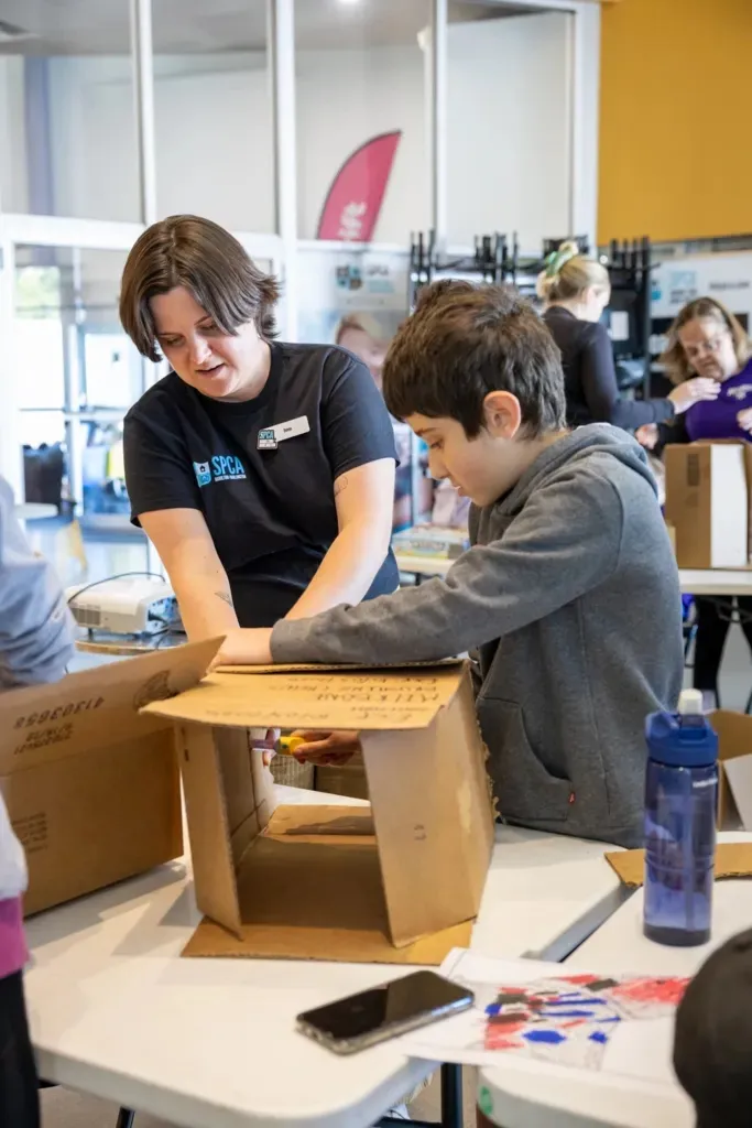 A woman and a boy assembling a cardboard box together at a table in a classroom setting.