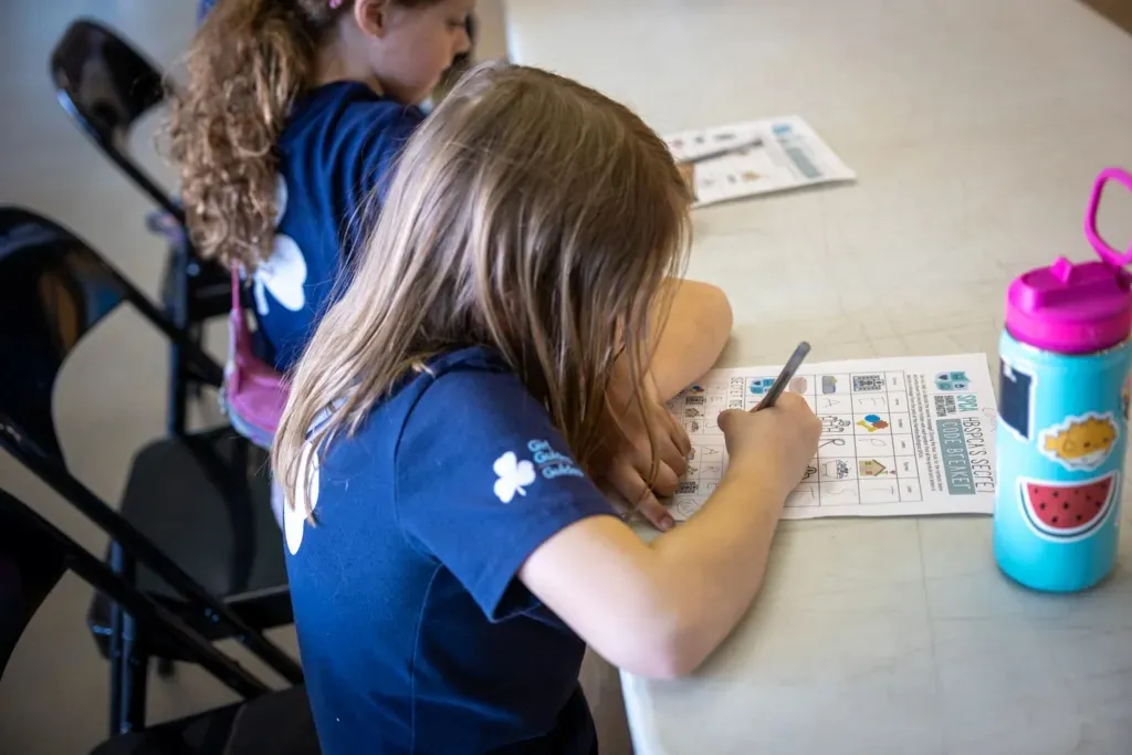Two children sitting at a table working on educational worksheets, with one child writing and a decorated water bottle nearby.
