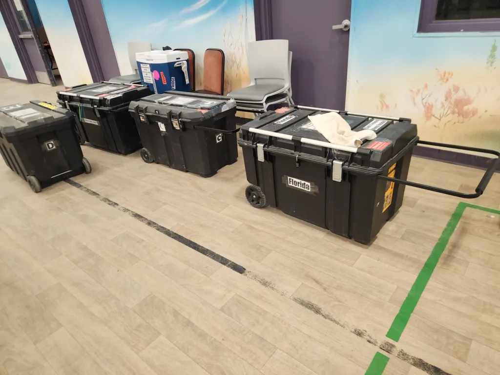 Four large black plastic storage containers on wheels lined up in a room with wooden flooring and painted walls.