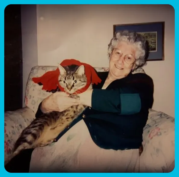 Elderly woman with glasses sitting on a floral armchair, holding a tabby cat wearing a red scarf.
