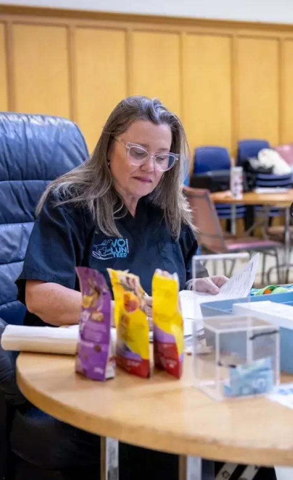Woman wearing glasses and a volunteer shirt sitting at a table reviewing papers with packaged snacks and clear donation boxes in front.