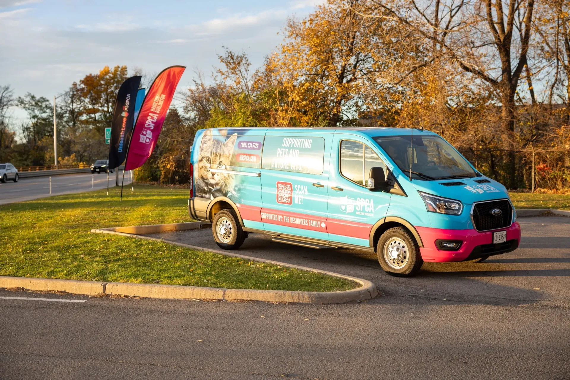 Blue and pink SPCA animal protection van parked in a lot with flags and autumn trees in the background.