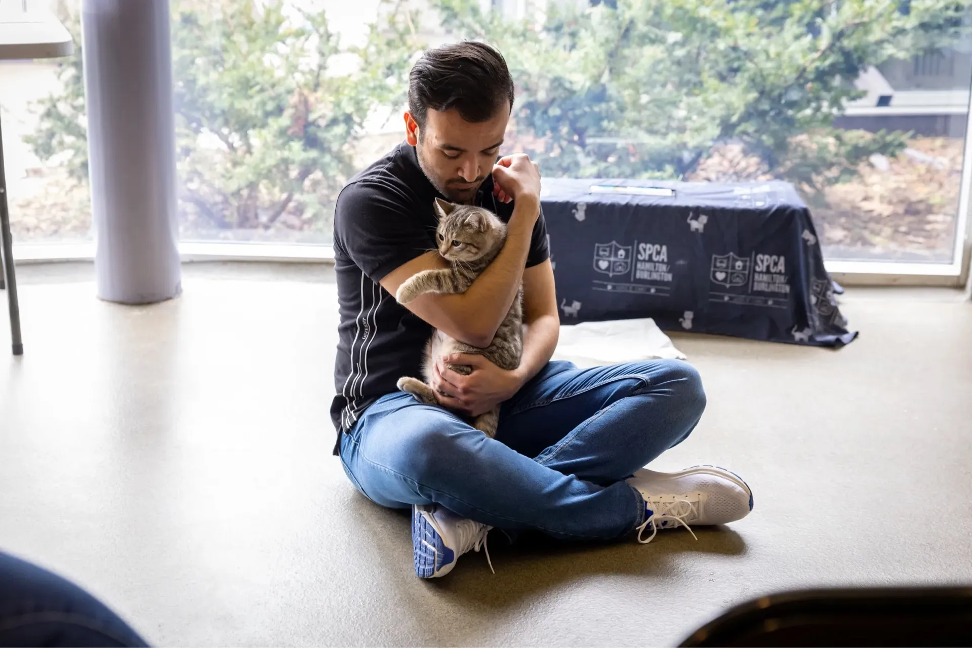 Man sitting cross-legged on the floor indoors, hugging a tabby cat close to his chest.