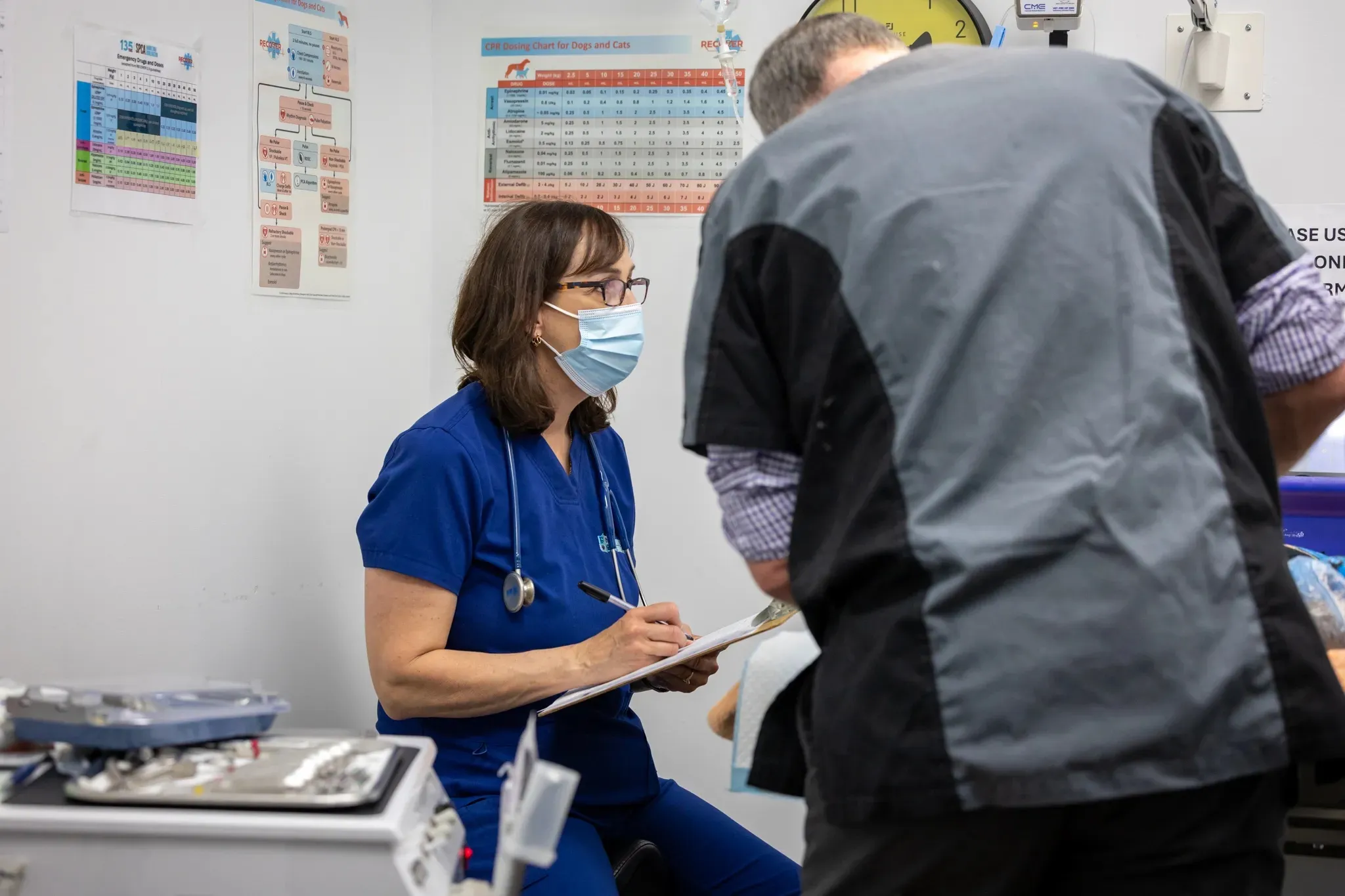 Masked female nurse in blue scrubs writing on a clipboard during a medical exam in a clinic room.