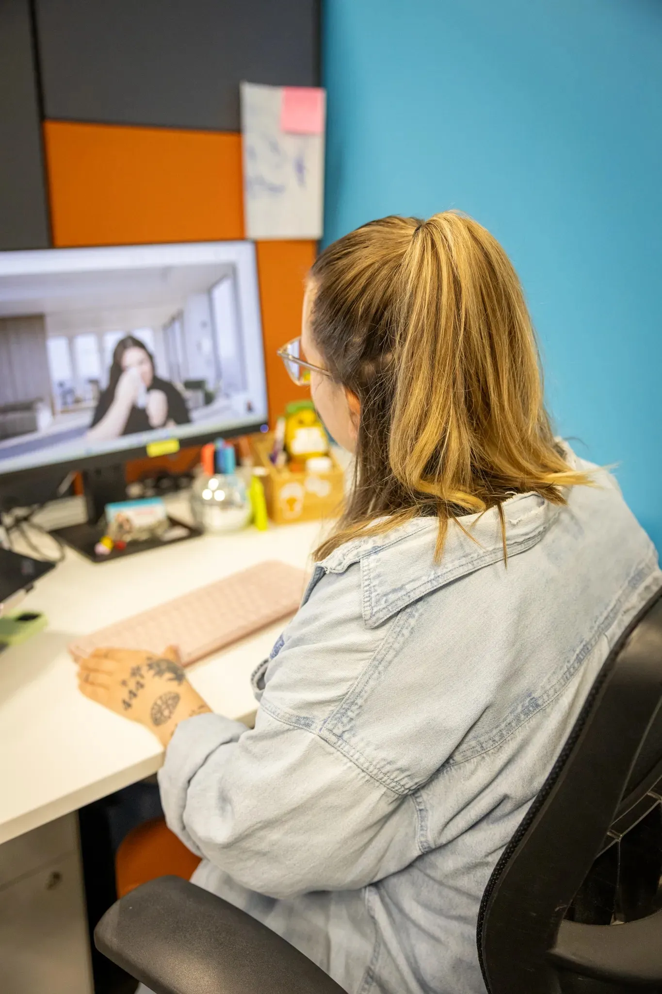 Person with a ponytail and denim jacket seated at desk, viewing a blurred video call on a computer screen.