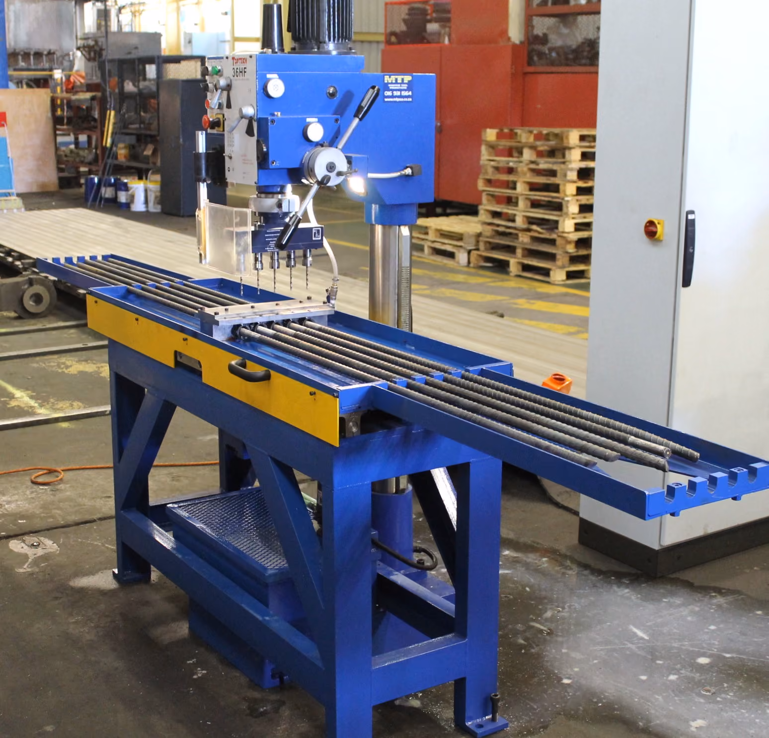 Blue multi-spindle drilling machine with metal rods set in an industrial workshop.