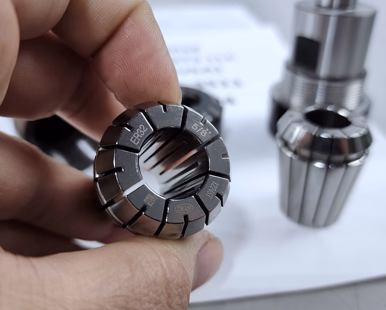 Hand holding metal ER32 collet marked 5/8 with other collets blurred on a white surface.