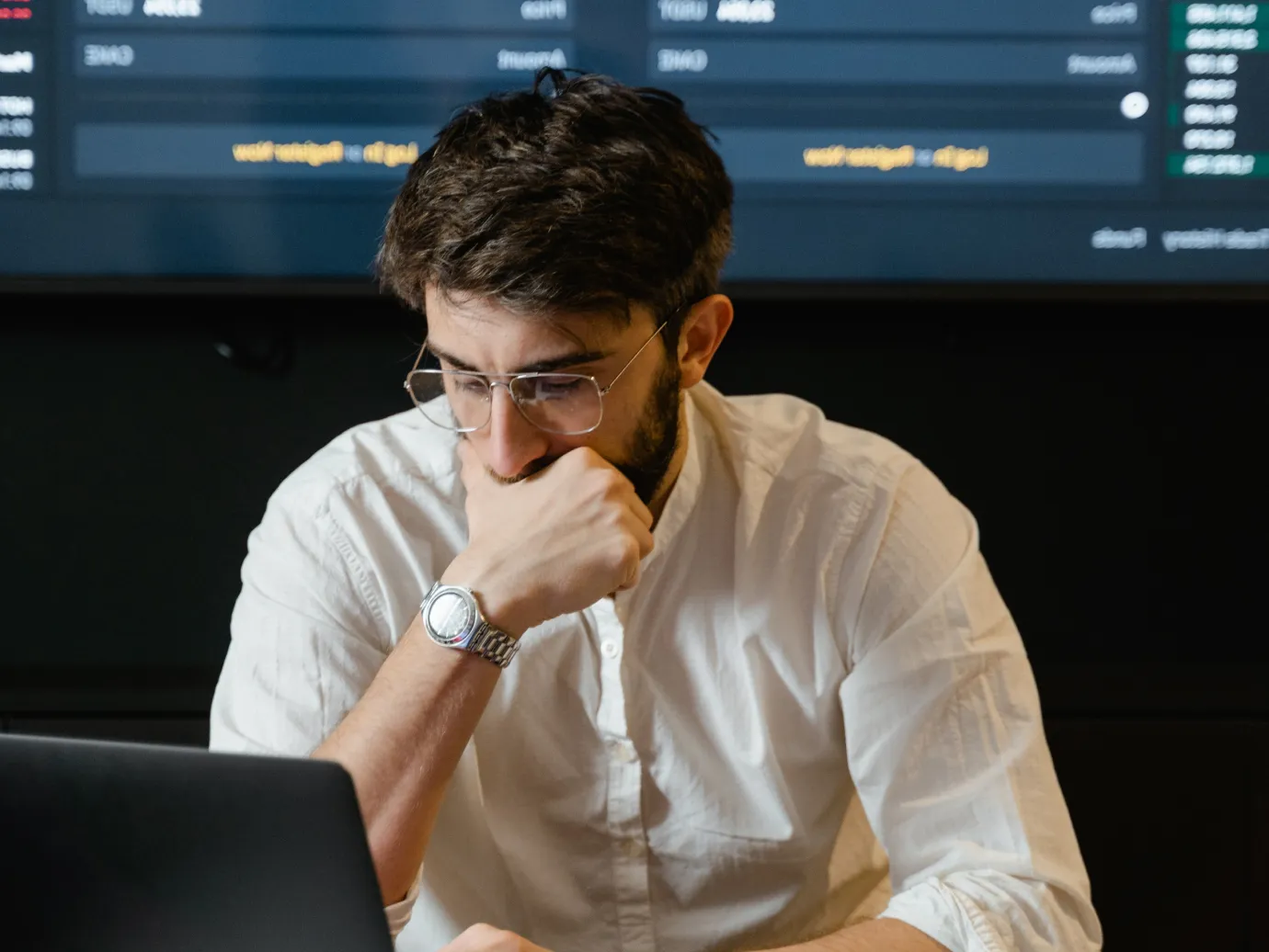 Man with glasses and a wristwatch, wearing a white shirt, looking thoughtfully at a laptop with a data screen in the background.