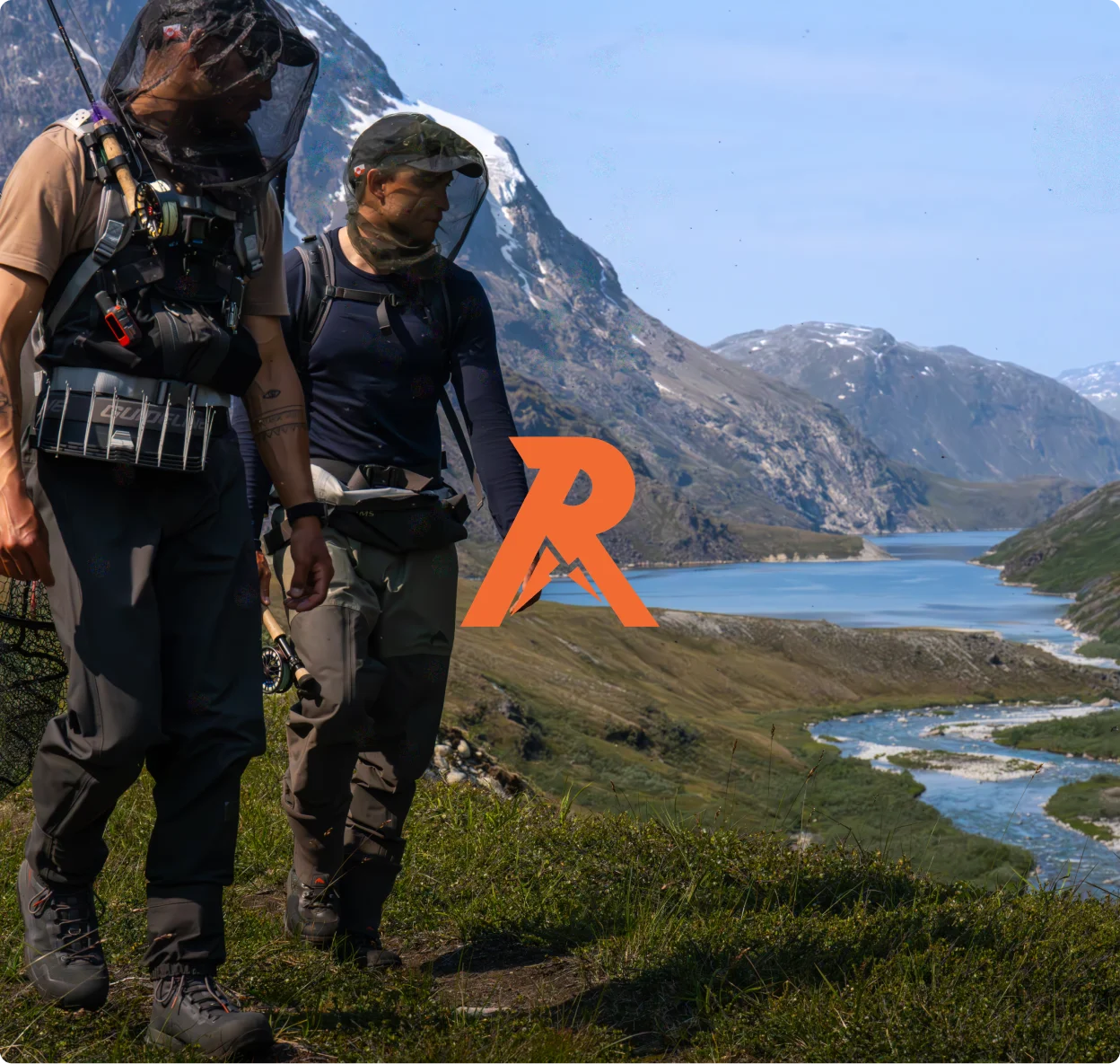 Two men wearing fishing gear walking on a trail by a river with mountains in the background.