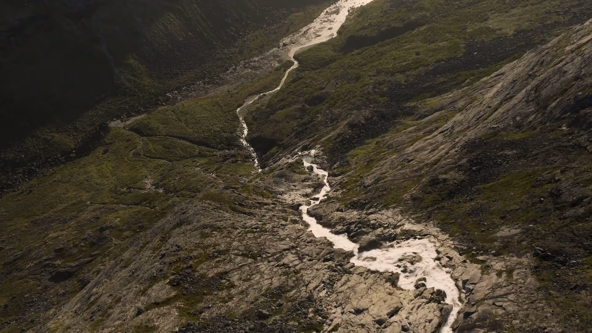 A narrow stream flows down a rocky hillside with patches of green grass.
