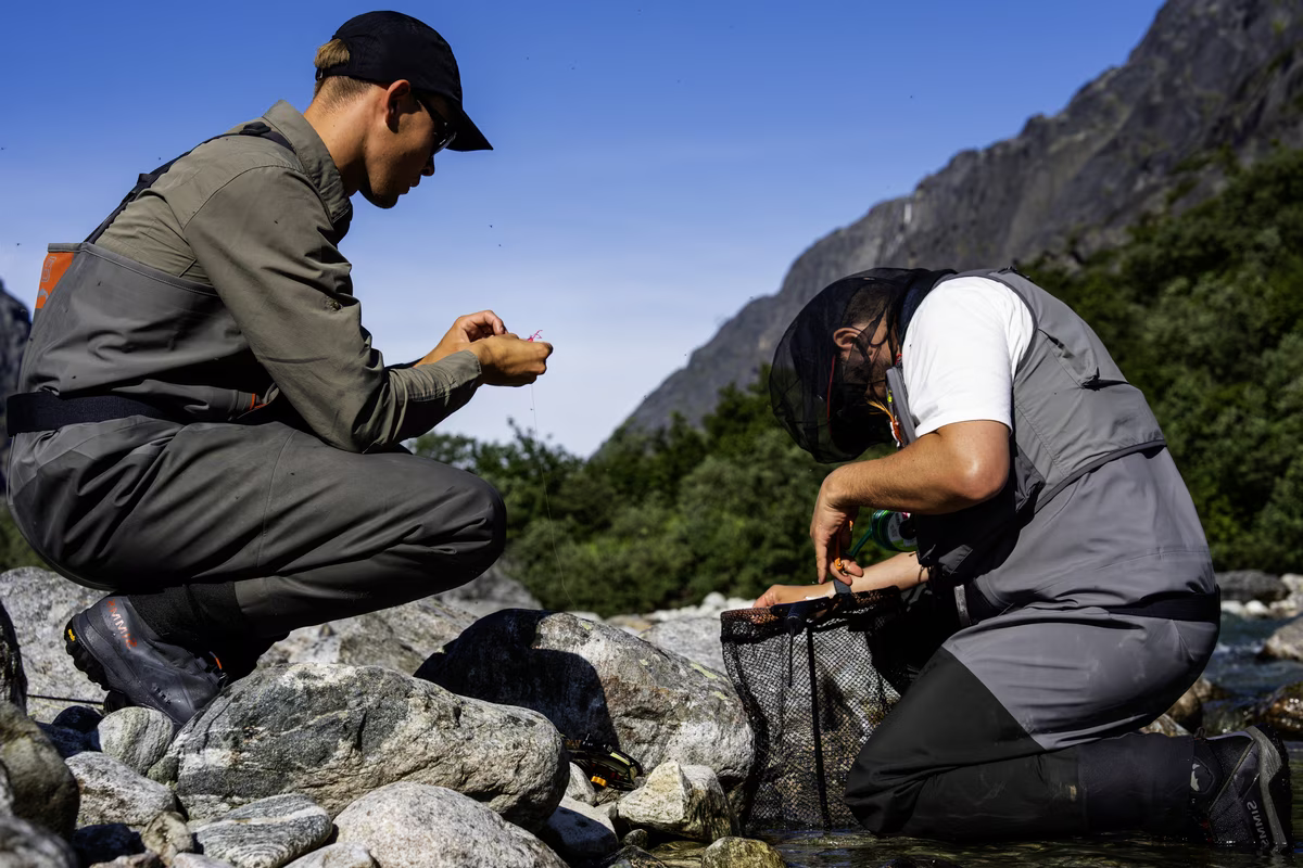 Two men in fishing gear preparing equipment by a rocky stream with mountains in the background.