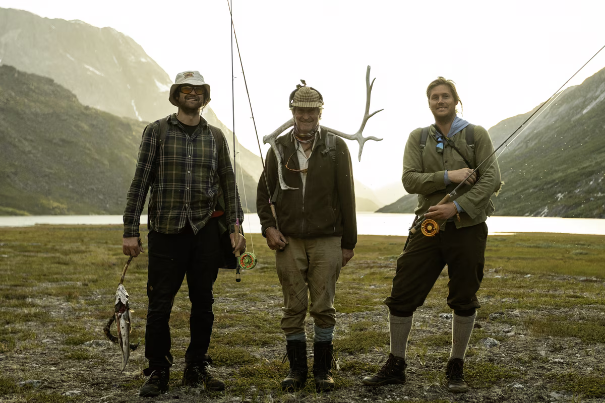 Three men holding fishing rods and a catch, standing on grassy land near a lake and mountains.