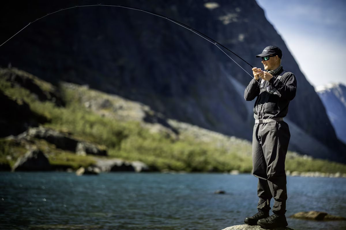 Man wearing sunglasses and cap fly fishing in a river with mountains in the background.