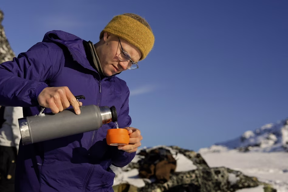 Person in yellow hat and purple jacket pouring drink from thermos into orange cup outdoors near snowy mountains.