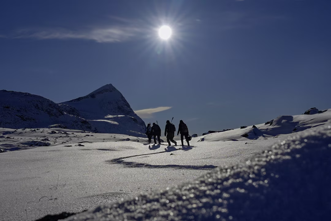 Four hikers trek across a sunlit snowy mountain landscape with a clear blue sky.