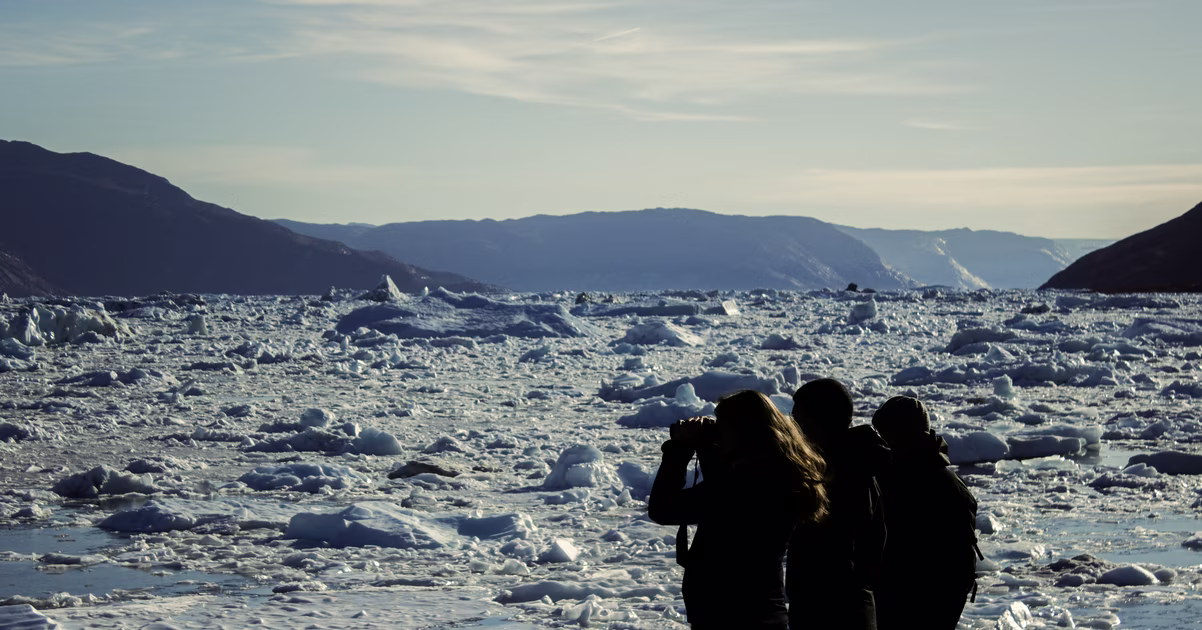 Three people silhouetted against a frozen sea with ice chunks and distant mountains at dusk.