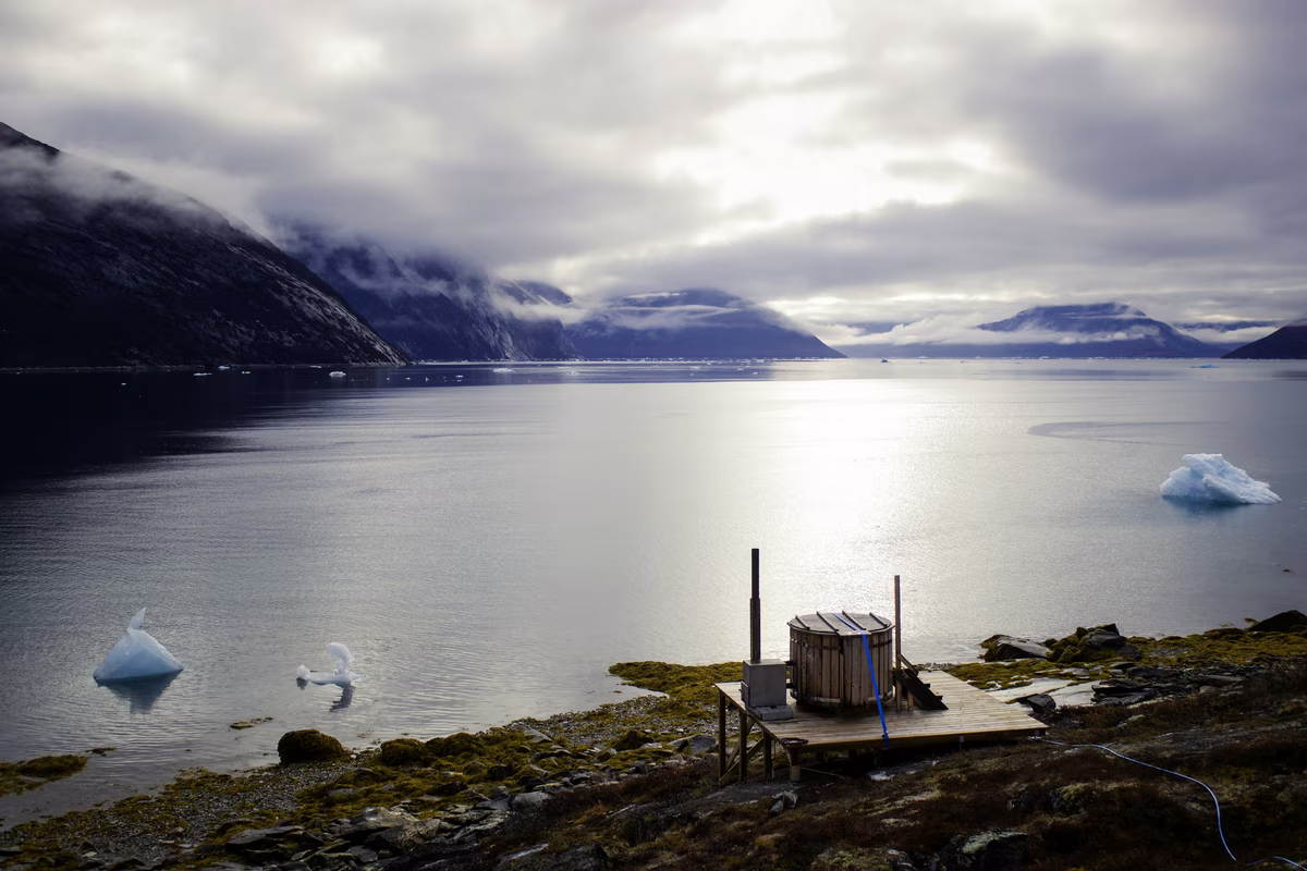 Wooden hot tub on deck near rocky shore with iceberg-filled fjord and misty mountains under cloudy sky.