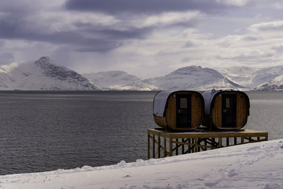 Two small arched cabins on stilts by a snowy fjord with mountains and cloudy sky in the background.