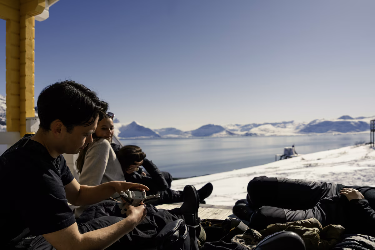 Group relaxing outdoors near snowy fjord under clear blue sky with mountains in the background.