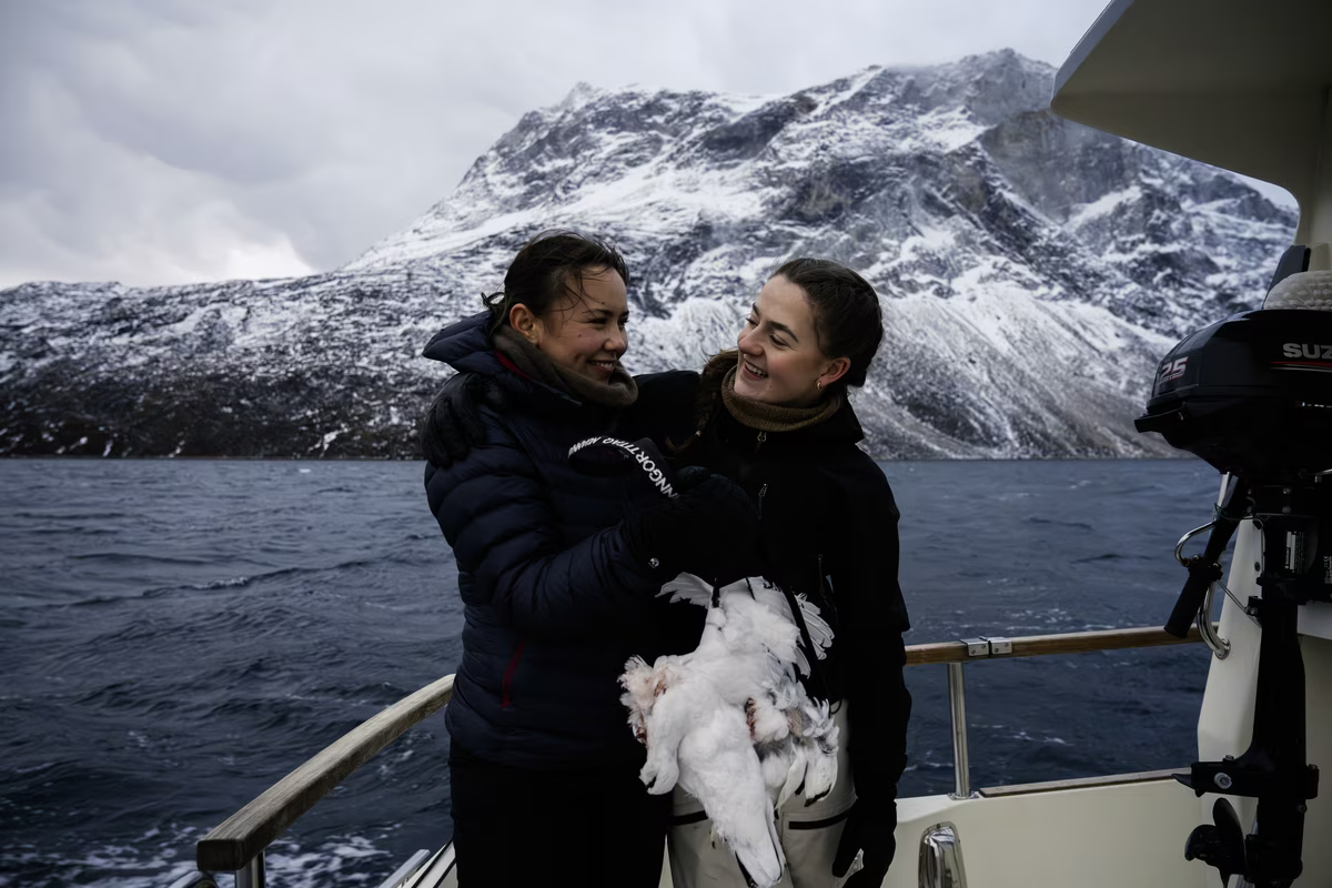 Two women smiling on boat holding a white bird with snowy mountain and sea in background.