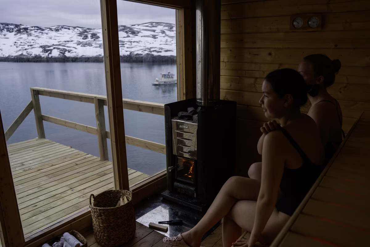 Two women in swimsuits sitting inside a wooden sauna by a stove, overlooking a snowy lake with a boat.