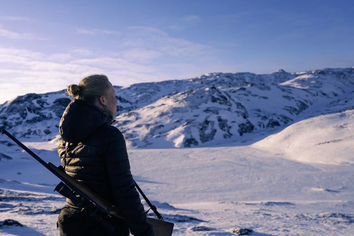 Person in a black jacket with a rifle on their back looking at snowy mountains under a clear sky.