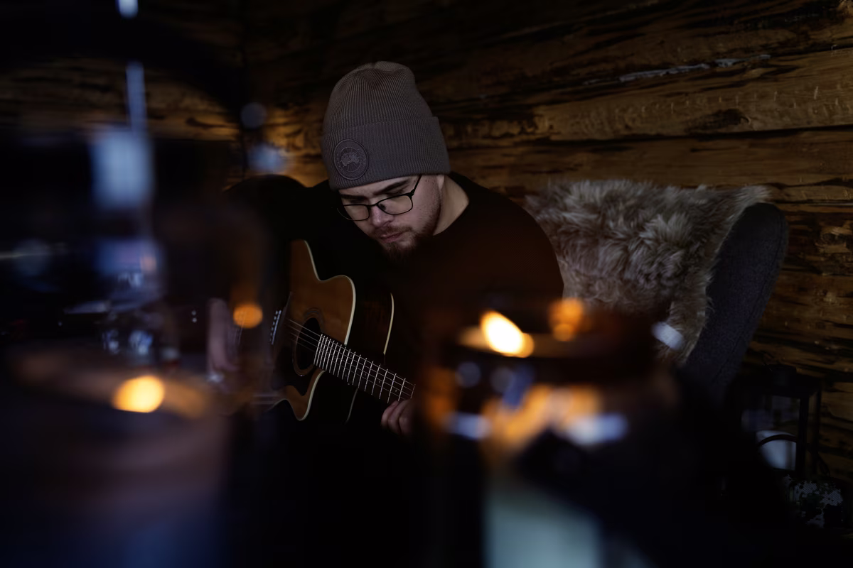 Man in glasses and beanie playing acoustic guitar in a dimly lit wooden cabin.