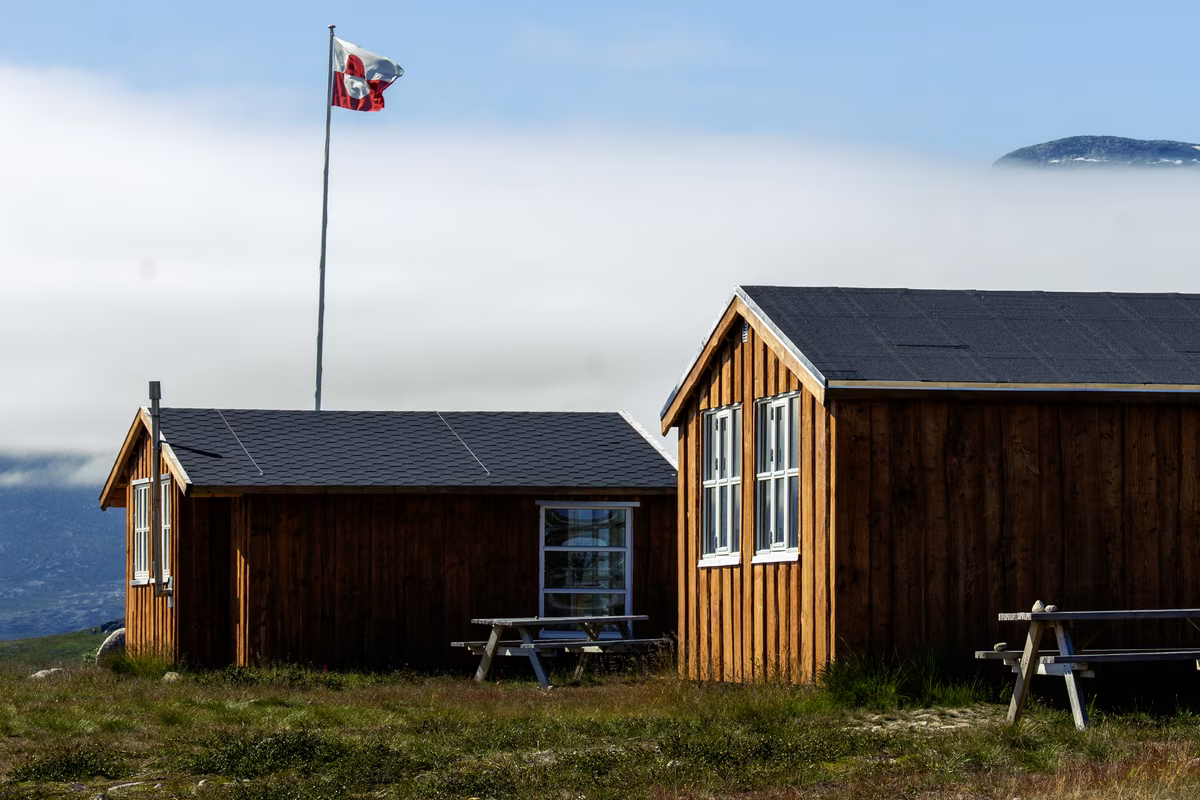 Two wooden cabins with picnic tables on grassy terrain under a blue sky with a flagpole flying a red and white flag.