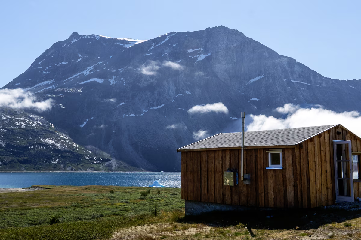 Wooden cabin near a lake with a large rocky mountain and scattered clouds in the background.
