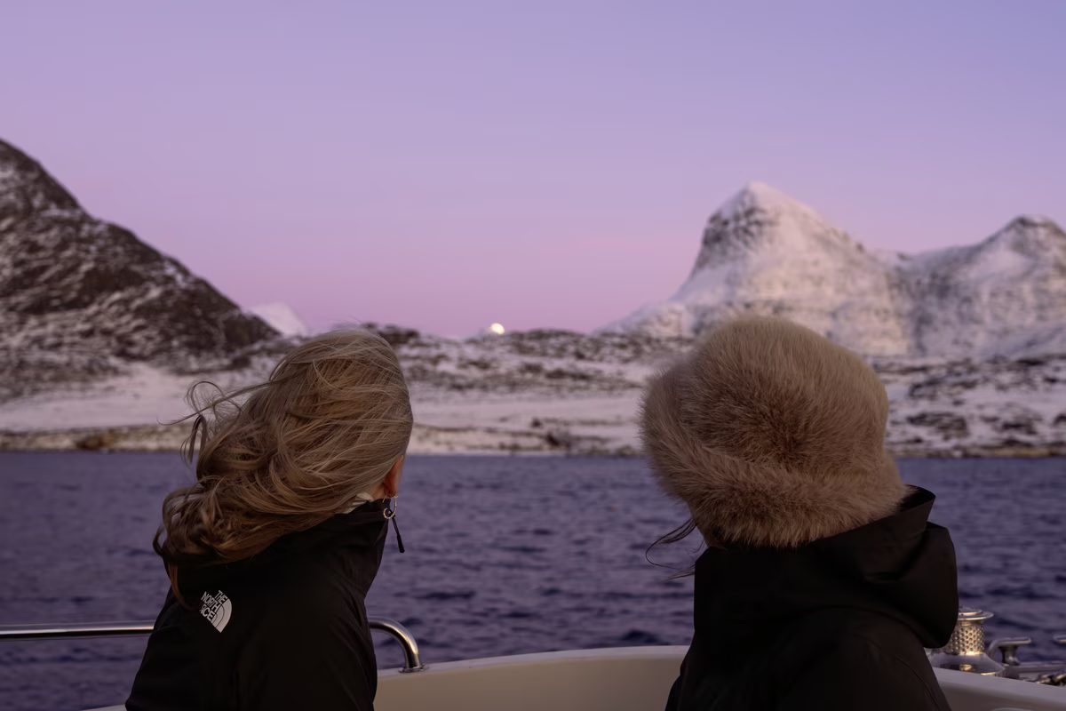 Two people in warm jackets looking at snowy mountains across a body of water during twilight.