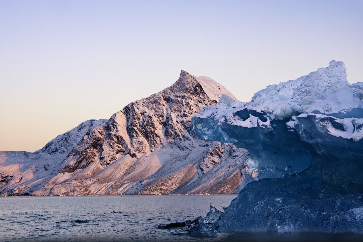 Snow-covered mountain behind a large, translucent blue iceberg in calm water at sunset.
