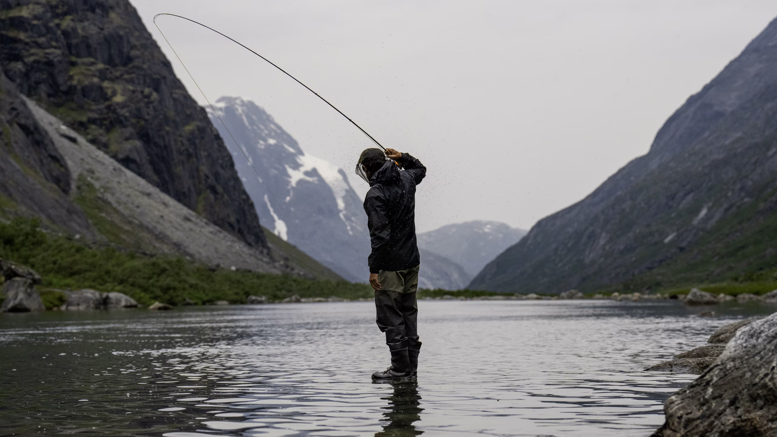 Person fly fishing in a river surrounded by tall mountains with snow patches under gray sky.